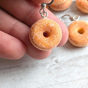 Könnte beinhalten: Ein Paar Donut-Ohrringe mit Zuckerguss. Die Donuts sind hellbraun und haben ein Loch in der Mitte. Die Ohrringe sind aus Silber und haben einen Hakenverschluss.
