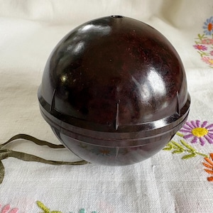 Puede incluir: Un objeto esférico de color marrón oscuro con un acabado brillante. El objeto se compone de dos partes, una superior y otra inferior, con líneas verticales. El objeto está sobre una superficie blanca con bordados florales.