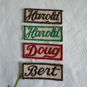 May include: Four rectangular embroidered name patches on a white fabric background. The patches feature the names Harold, Harold, Doug, and Bert in cursive script. The borders and text are in brown, green, and red thread on a beige background.