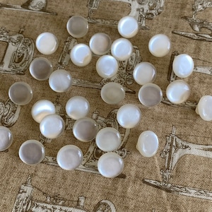 May include: A collection of white pearl-like buttons, scattered on a fabric background with a sewing machine design.