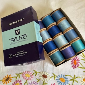 Op de afbeelding: Een doos met Dewhurst Sylko gemerceriseerd katoengaren met twaalf houten klossen in verschillende blauwtinten. De donkerpaarse en lichtblauwe doos bevat de tekst Dewhurst, Sylko en English Sewing Ltd. De klossen zijn in rijen gerangschikt.