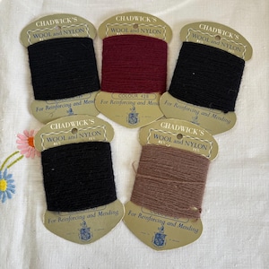 Może przedstawiać: Pięć szpulek nici Chadwick's Wool and Nylon w różnych kolorach, w tym czarnym, bordowym i brązowym. Każda szpulka ma papierową etykietę z napisem "For Reinforcing and Mending" i numerem koloru.