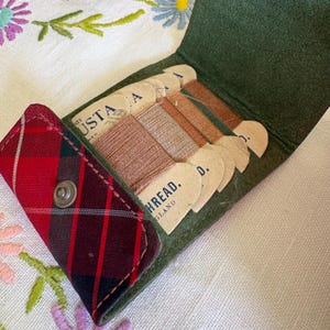 May include: A green leather case with a red and black plaid fabric flap featuring a metal snap closure. Inside, the case holds several cardboard spools wrapped in brown thread, each printed with the text "USTA" and "THREAD. ENGLAND".