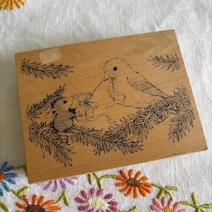 May include: A wooden block featuring a detailed black and white illustration of a bird offering a gift to a mouse. The bird is perched on a branch, and the mouse is holding the gift. The block has a light brown color.