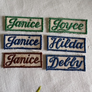 May include: Six rectangular embroidered name patches on a white fabric background. The patches feature the names Janice, Joyce, Janice, Hilda, Janice, and Debby in cursive script, with borders in green, blue, and brown matching the text colors.