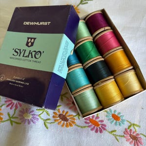 May include: A box of Dewhurst Sylko mercerised cotton thread containing twelve wooden spools in various colours including purple, pink, yellow, green, and blue. The dark blue and light blue box features the text Dewhurst Sylko and English Sewing Ltd.