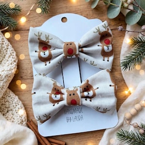 May include: Two fabric hair bows featuring a repeating pattern of brown reindeer with red noses on a light beige background. The bows are attached to a white display card with the text "HANDMADE BY LAURA" printed in black on the lower centre.