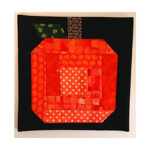 Op de afbeelding: Een rood en oranje patchwork quiltblok in de vorm van een pompoen. Het blok is gemaakt van vierkanten en rechthoeken van verschillende stoffen, sommige met een pompoenprint, sommige met stippen en sommige met een effen kleur. Het blok is omgeven door een zwarte rand.