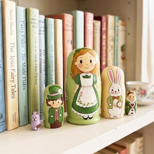 May include: A collection of hand-painted wooden nesting dolls, featuring characters from Alice in Wonderland. The dolls are in various sizes and colors, including green, white, and pink, and are displayed on a white shelf with books in the background.