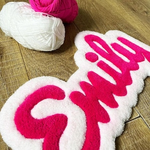 May include: A tufted rug featuring the name Emily in bright pink yarn against a white background, accompanied by one ball of hot pink yarn and one ball of white yarn. The rug has a soft, textured surface with a distinct, raised script lettering.