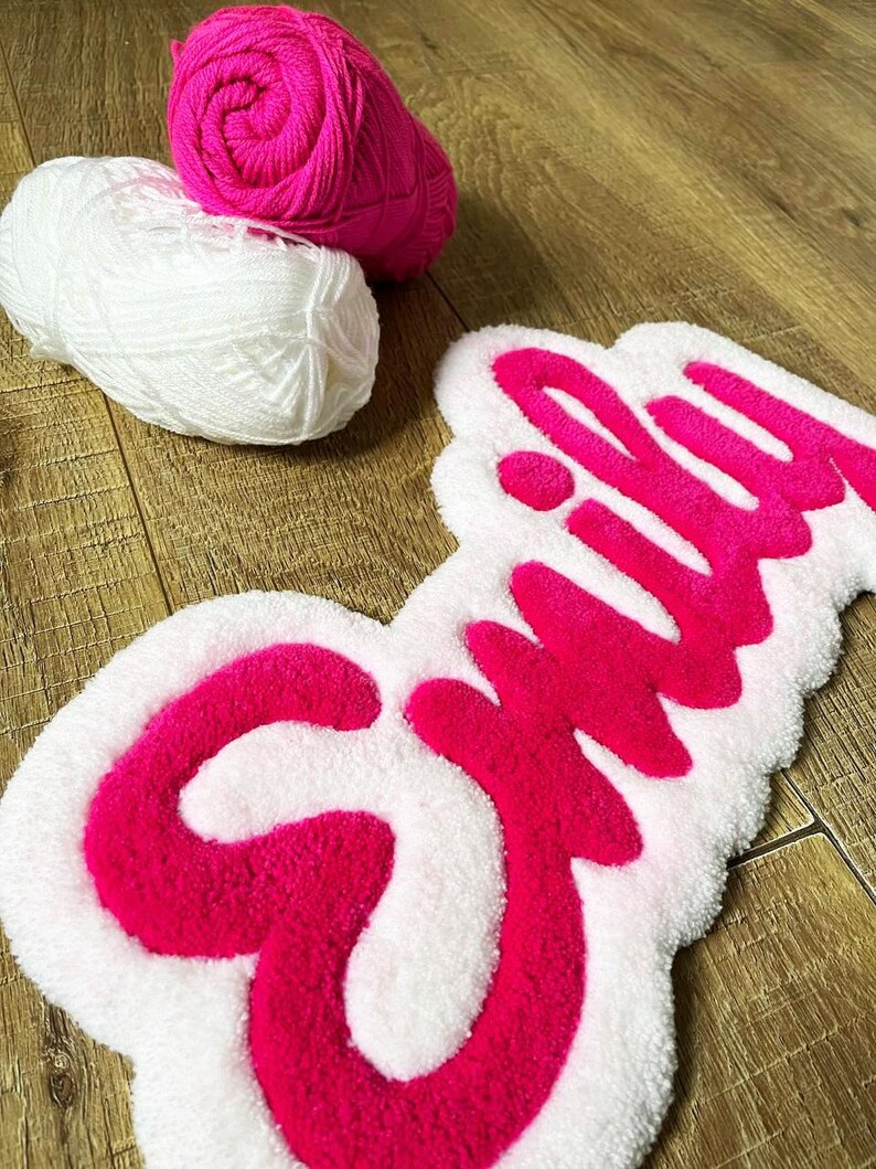 May include: A tufted rug featuring the name Emily in bright pink yarn against a white background, accompanied by one ball of hot pink yarn and one ball of white yarn. The rug has a soft, textured surface with a distinct, raised script lettering.