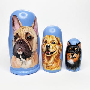 Pode incluir: Conjunto de três bonecas russas pintadas à mão com retratos de cães. As bonecas retratam um Buldogue Francês, um Golden Retriever e um cão pequeno preto e castanho, sobre um fundo azul claro.