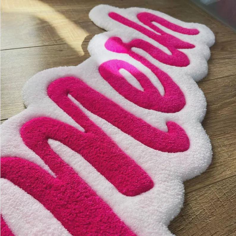 May include: A tufted rug featuring a white background with the word "MOM" written in large, bold, bright pink cursive lettering. The rug has a soft, textured pile surface and follows the organic, rounded shape of the script lettering.