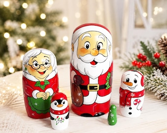 May include: A set of five wooden nesting dolls featuring a Santa Claus, a Mrs. Claus with glasses, a snowman with a red scarf, a small penguin with string lights, and a tiny solid green figure, all painted in festive holiday colours.