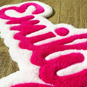 May include: A tufted rug featuring a white background with the word "Smile" written in bright pink yarn. The text is rendered in a cursive script font with a heart shape integrated into the letter S, showcasing a thick, textured pile surface.