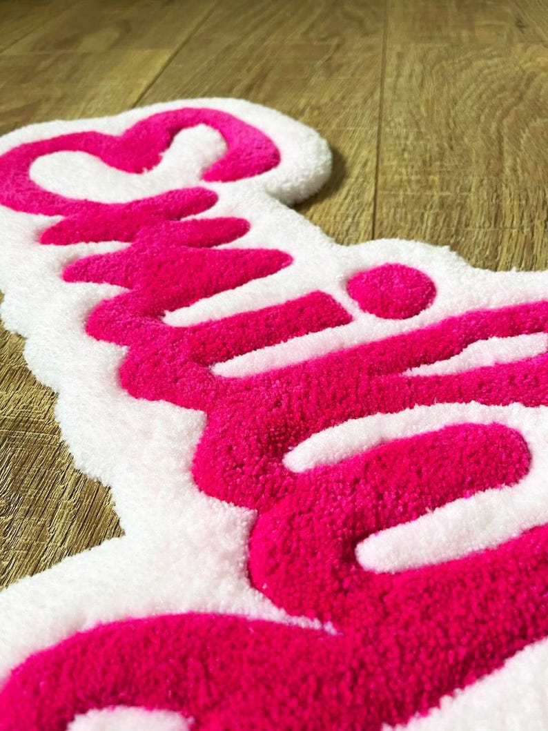 May include: A tufted rug featuring a white background with the word "Smile" written in bright pink yarn. The text is rendered in a cursive script font with a heart shape integrated into the letter S, showcasing a thick, textured pile surface.