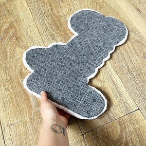 May include: A shaped rug with a gray, textured, non-slip backing featuring a repeating pattern of small black dots. The rug has a white, fuzzy, plush border that outlines its irregular, organic shape, providing a soft contrast to the gray surface.
