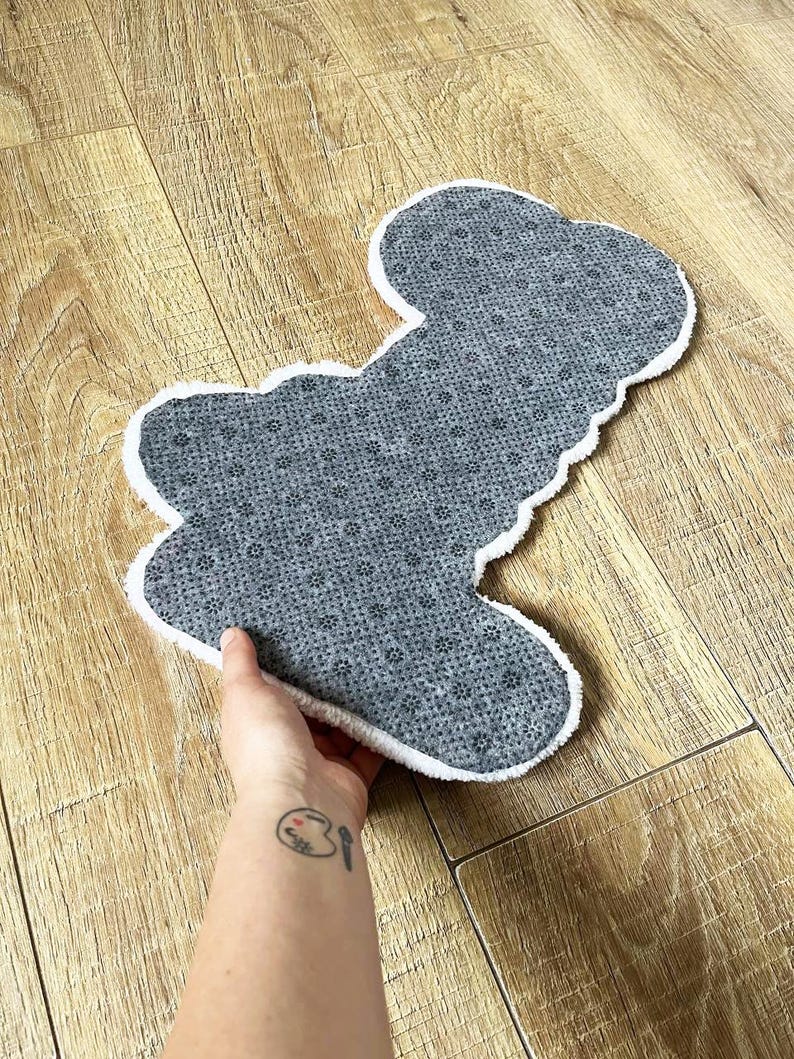 May include: A shaped rug with a gray, textured, non-slip backing featuring a repeating pattern of small black dots. The rug has a white, fuzzy, plush border that outlines its irregular, organic shape, providing a soft contrast to the gray surface.