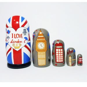 Pode incluir: Um conjunto de cinco bonecas russas, ou bonecas matrioska, pintadas com desenhos temáticos de Londres. A boneca maior é vermelha, branca e azul com as palavras "I Love London" em um coração. As outras bonecas apresentam uma torre do relógio Big Ben, uma cabine telefônica vermelha, um ônibus de Londres e uma coroa.