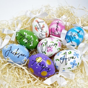 May include: A collection of hand-painted Easter egg ornaments in various colors, including green, blue, purple, and white. Each egg features a different name in black script and is adorned with floral or patterned designs. White ribbons are attached for hanging.