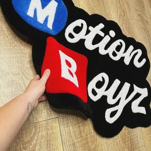 May include: A custom tufted rug featuring a black background with the text Motion Boyr. The letter M is white on a blue oval, and the letter B is white on a red square. The remaining letters are white and cursive, creating a bold, irregular shape.