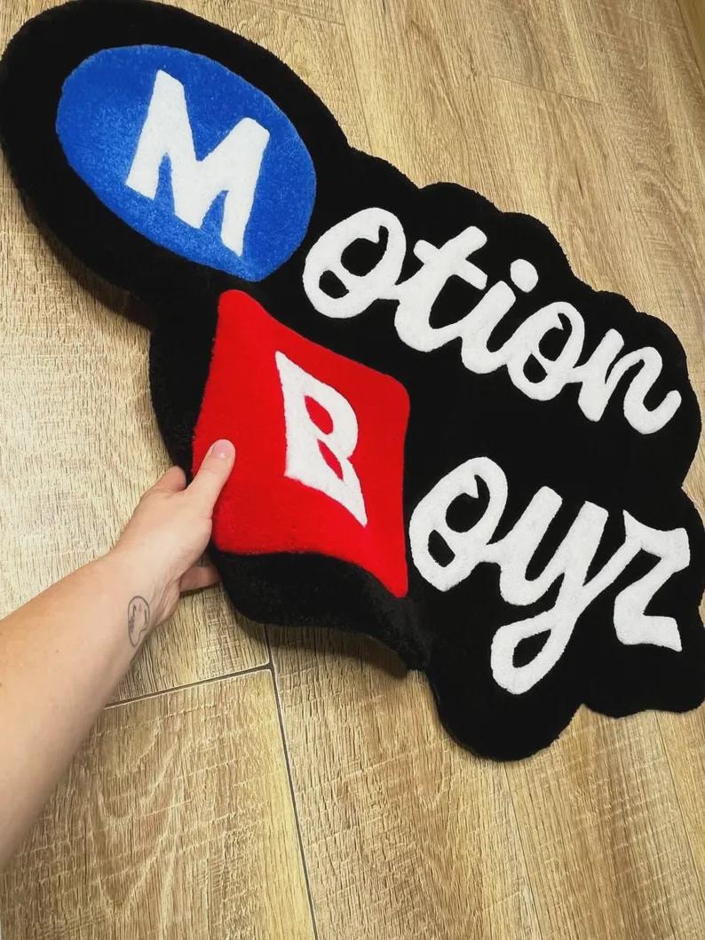 May include: A custom tufted rug featuring a black background with the text Motion Boyr. The letter M is white on a blue oval, and the letter B is white on a red square. The remaining letters are white and cursive, creating a bold, irregular shape.