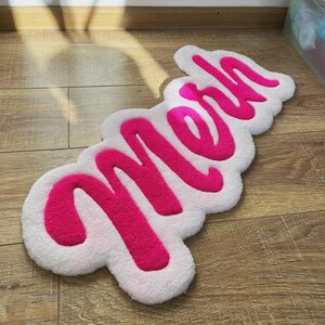 May include: A tufted rug featuring the word "meh" in bright pink yarn, set against a white, cloud-shaped background. The rug has a soft, textured pile and follows the cursive lettering of the word, creating a distinct, irregular shape.