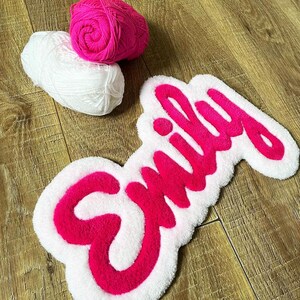 May include: A tufted rug featuring the name Emily in bright pink yarn with a white border, accompanied by one ball of bright pink yarn and one ball of white yarn. The rug has a soft, textured surface and a cursive script font style.