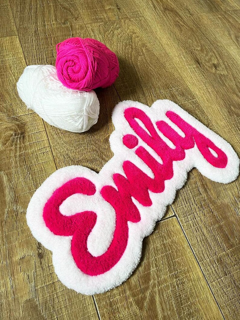 May include: A tufted rug featuring the name Emily in bright pink yarn with a white border, accompanied by one ball of bright pink yarn and one ball of white yarn. The rug has a soft, textured surface and a cursive script font style.