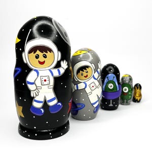 May include: A set of six nesting dolls with an outer space theme. The largest doll is black with a white astronaut design, stars, and a yellow star. The other dolls feature astronauts and aliens in various colors, including gray, green, and black.