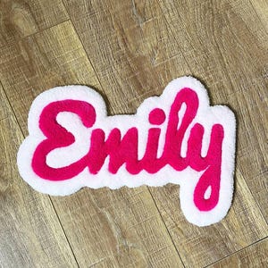 May include: A custom tufted rug featuring the name Emily in bright pink cursive lettering. The text is outlined with a thick, white, cloud-shaped border that follows the contours of the letters, creating a soft, textured, and layered appearance.