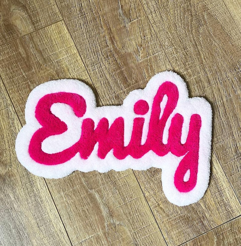 May include: A custom tufted rug featuring the name Emily in bright pink cursive lettering. The text is outlined with a thick, white, cloud-shaped border that follows the contours of the letters, creating a soft, textured, and layered appearance.