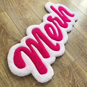 May include: A tufted rug featuring the word "Meh" in bright pink yarn, set against a white, cloud-shaped background. The text is written in a bold, cursive script, and the entire piece has a soft, textured pile surface with rounded edges.