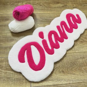 May include: A white tufted rug featuring the name Diana in bright pink cursive script, accompanied by one ball of bright pink yarn and one ball of white yarn, showcasing the texture and color palette used for the custom handmade textile piece.