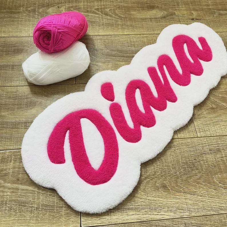 May include: A white tufted rug featuring the name Diana in bright pink cursive script, accompanied by one ball of bright pink yarn and one ball of white yarn, showcasing the texture and color palette used for the custom handmade textile piece.