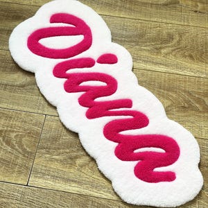 May include: A tufted rug with a white base and a bright pink script font spelling out the name Diana. The rug features a soft, plush texture with a rounded, organic shape that follows the contours of the lettering in a bold, raised design.
