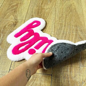 May include: A tufted rug featuring the text "hm" in bright pink yarn against a white background. The rug has an irregular, cloud-like shape with a soft, textured pile on the front and a dark gray, non-slip backing visible on the folded edge.