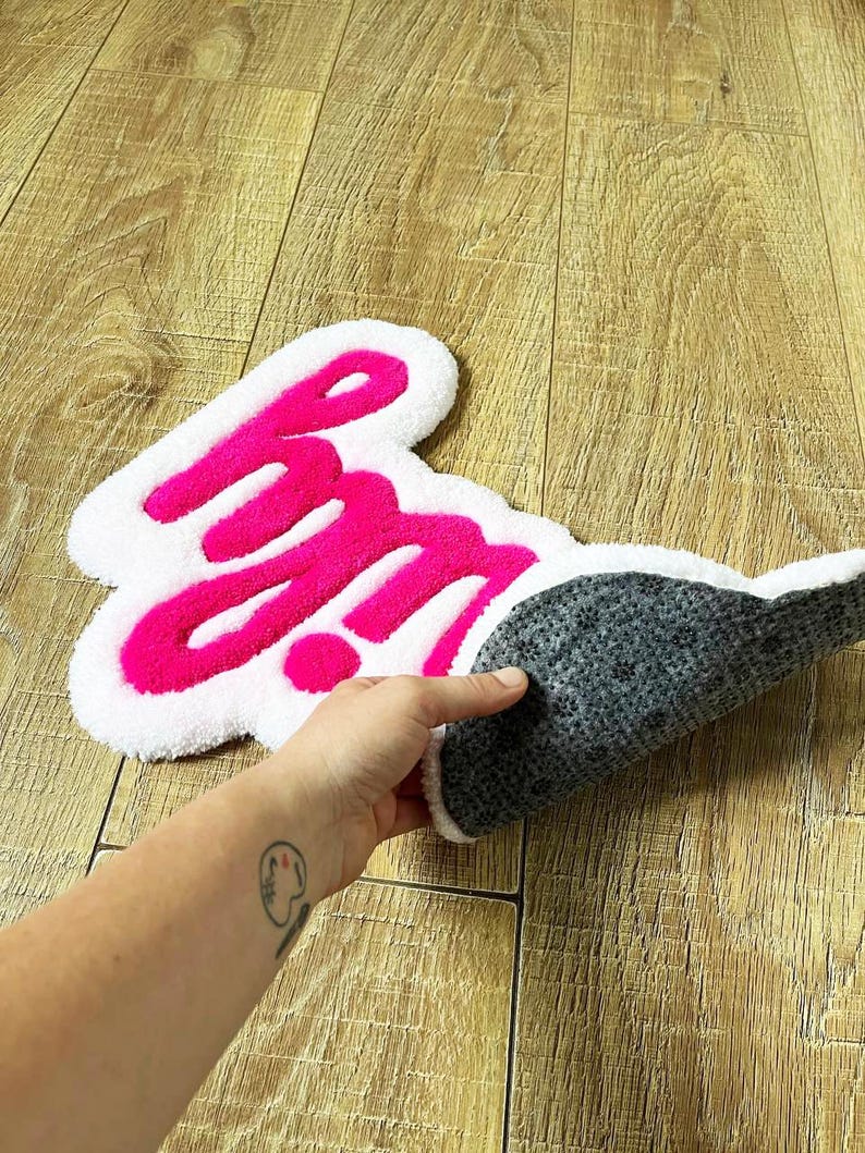 May include: A tufted rug featuring the text "hm" in bright pink yarn against a white background. The rug has an irregular, cloud-like shape with a soft, textured pile on the front and a dark gray, non-slip backing visible on the folded edge.