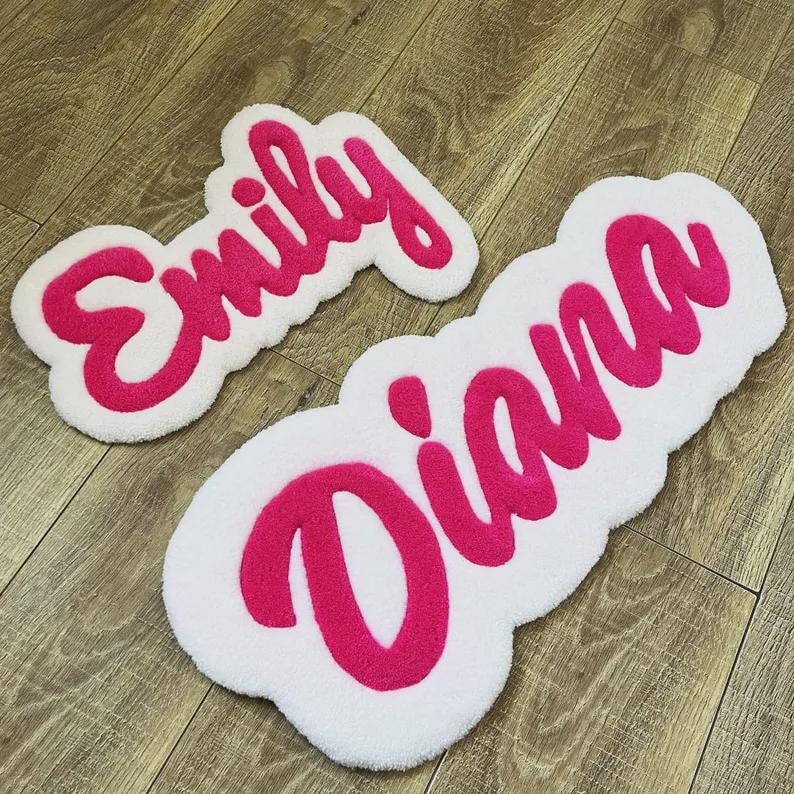 May include: Two custom tufted rugs with white backgrounds and bright pink cursive lettering. One rug displays the name Emily, and the larger rug displays the name Diana. Both rugs feature a soft, textured pile and rounded, organic edges.