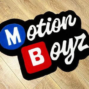 May include: A custom tufted rug featuring the text Motion Boyz. The letter M is white inside a blue circle, the letter B is white inside a red square, and the remaining letters are white script on a black background with a thick black border.