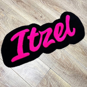 May include: A custom tufted rug featuring the name Itzel in bright pink cursive lettering against a solid black background. The rug has an irregular, rounded shape that follows the contour of the text, creating a bold and high-contrast design.