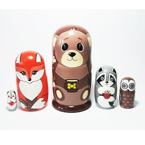May include: A set of five wooden nesting dolls, or matryoshka dolls, featuring different woodland animals. The largest doll is a brown bear holding a cup of honey, followed by a red fox, a gray raccoon holding a raspberry, and a brown owl. The smallest doll is a gray squirrel holding a red apple.