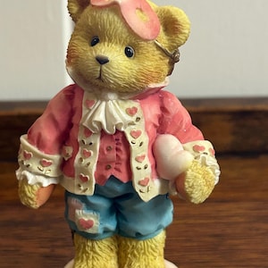 May include: A decorative teddy bear figurine. The bear is dressed in a pink jacket with heart details, blue shorts, and a pink headpiece. It holds a heart-shaped object and stands on a small, white base. The bear's fur is yellow.