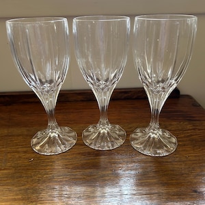 Puede incluir: Tres copas de vino transparentes con un diseño acanalado en la copa. Las copas tienen una apariencia delicada y elegante, con una base redondeada y un tallo delgado. Están dispuestas sobre una superficie de madera oscura.