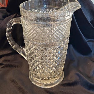 May include: Clear glass pitcher with a diamond pattern design. The pitcher has a handle and a spout for pouring. The pitcher is made of clear glass and is likely used for serving beverages.