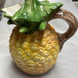 May include: A ceramic pitcher shaped like a pineapple. The pitcher has a textured yellow and orange body, a green leafy top, and a brown handle. The pitcher is designed to resemble a pineapple fruit.