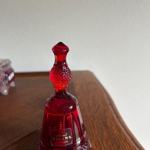 Puede incluir: Una campana de cristal rojo rubí con un asa texturizada y una base con diseño de diamantes. La campana tiene una etiqueta que dice "VIKING MADE". La campana está sobre una superficie de madera.