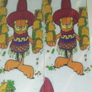 Puede incluir: Un par de calcetines blancos con una imagen de dibujos animados de Garfield usando un sombrero y sosteniendo una pila de tacos. Los calcetines tienen un esquema de color rojo, amarillo y verde.