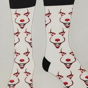 May include: A pair of white crew socks featuring a repeating pattern of a clown face with red accents, including a red nose, red lips, and sharp teeth. The socks have a solid black cuff at the top and a solid black toe section at the bottom.