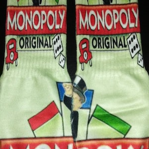 May include: A pair of green and white socks with a Monopoly board game design. The socks feature the Monopoly man, dice, and the words "MONOPOLY ORIGINAL".